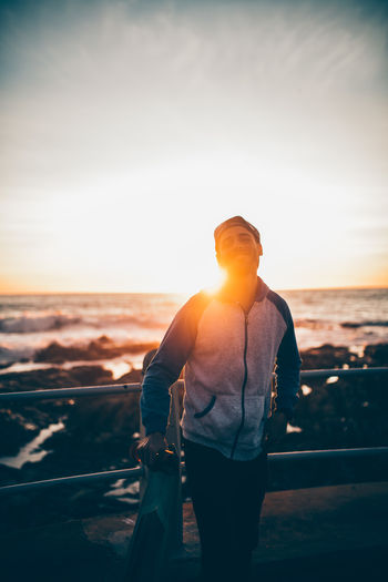 Back lit skateboarder standing with skateboard by railing against sky during sunset