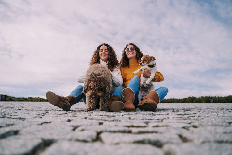 Low angle view of women holding dogs while sitting on land against sky