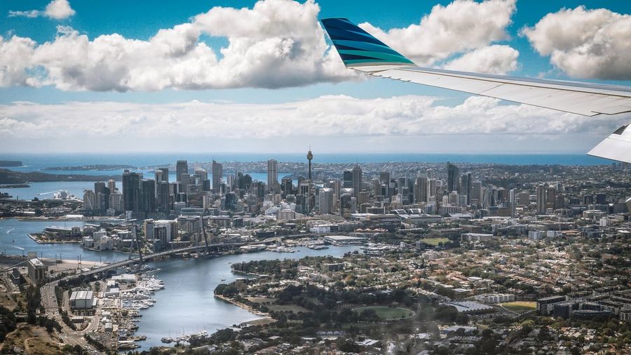 Cropped wing airplane flying above city against cloudy sky