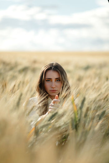 Young female with wavy hair looking at camera in countryside field under cloudy sky on blurred background