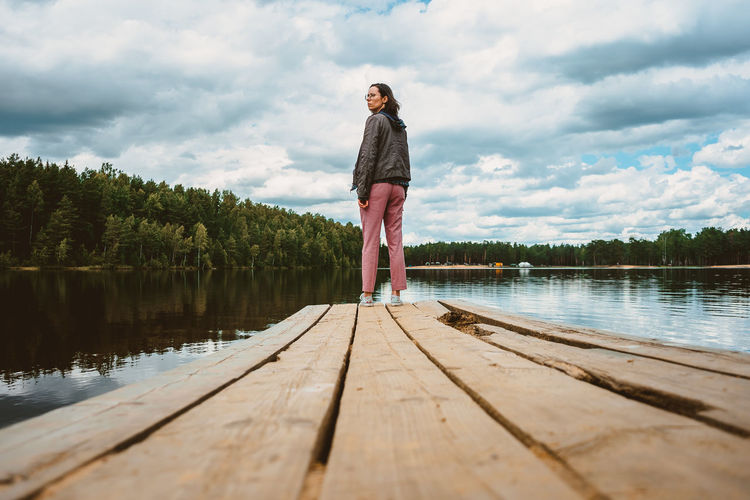 The tourist girl stands with her back on the wooden pier of the forest lake and turns around