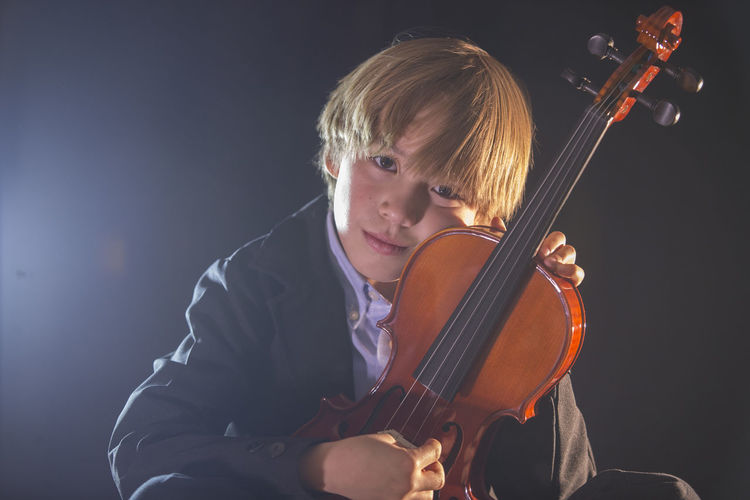 Boy holding violin while sitting against black background
