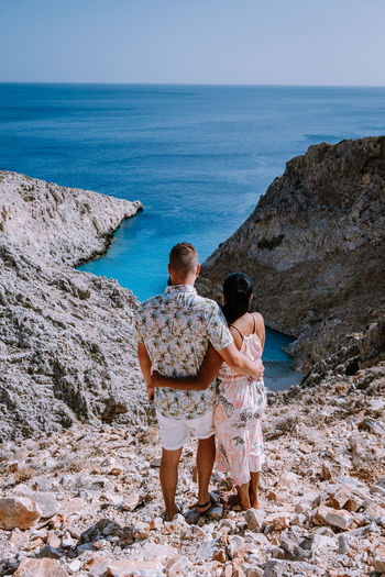 Rear view of couple standing on rock formation by sea against sky