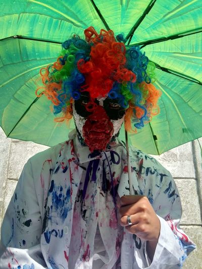 Young man in clown dressing with umbrella standing against wall