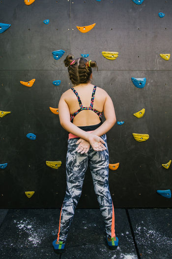 Back view of anonymous child in sportswear standing near wall with artificial rocks while training in gym and enjoying weekend