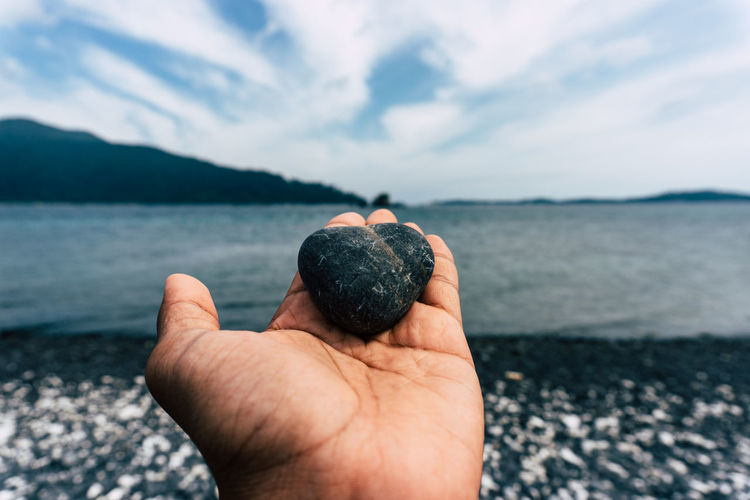 Cropped hand holding pebble at beach against sky
