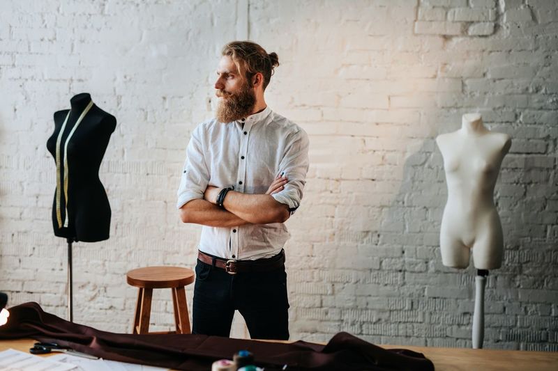 Fashion designer with arms crossed looking away while standing against brick wall in workshop
