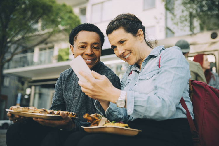 Smiling woman taking selfie with man while eating meal in city