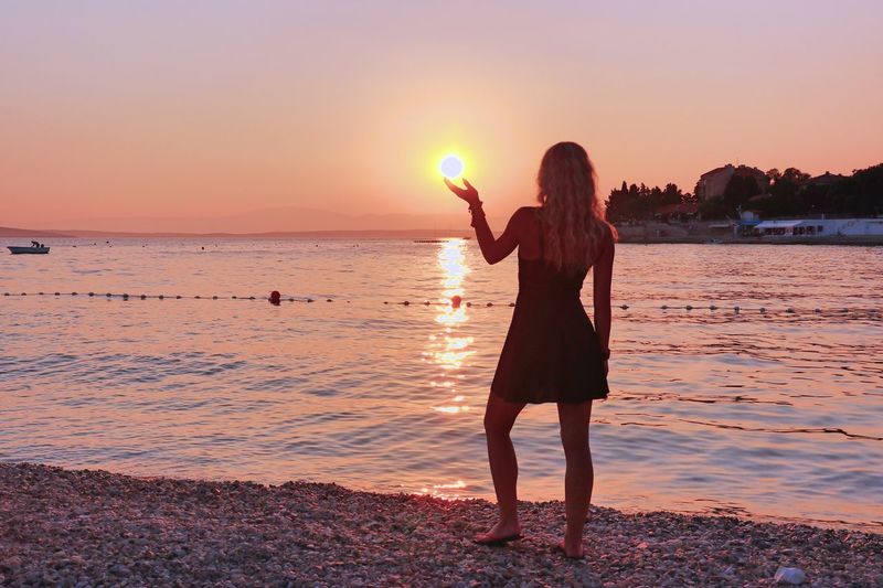 Optical illusion of woman holding sun while standing at beach against sky during sunset