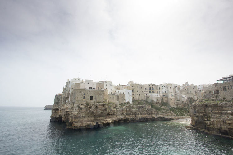 Buildings on cliff by sea at polignano a mare against cloudy sky