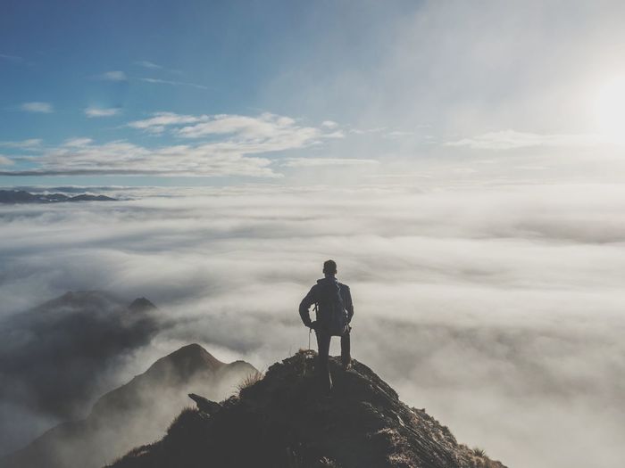 Rear view of young man with backpack standing on mountain against cloudy sky