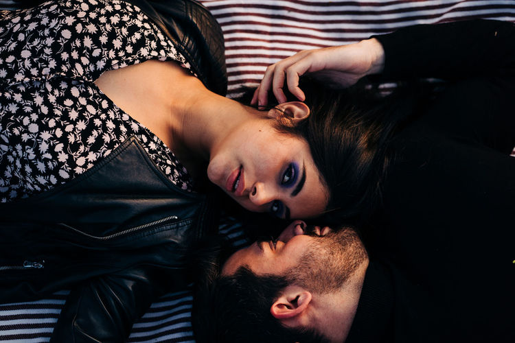 Top view of young ethnic woman with makeup in leather jacket lying near bearded male beloved on textile