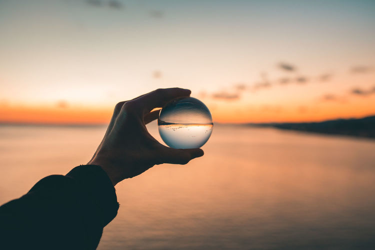 Cropped hand holding crystal ball against sky during sunset