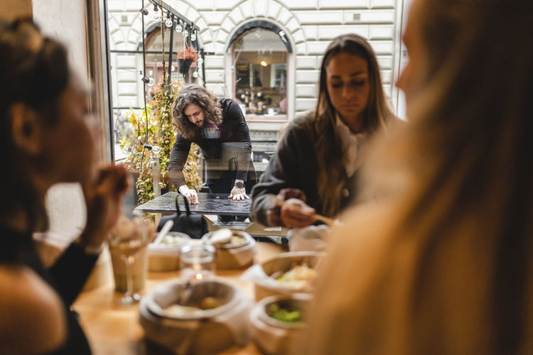 Multi-ethnic female friends having food in restaurant while waiter cleaning table outside