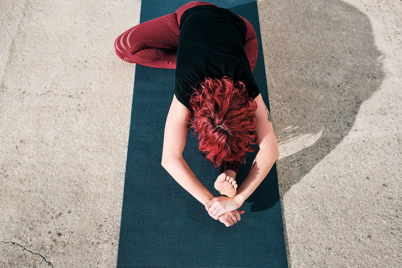 From above top view of unrecognizable flexible barefooted female athlete with red curly hair in activewear sitting practicing yoga on street among urban environment
