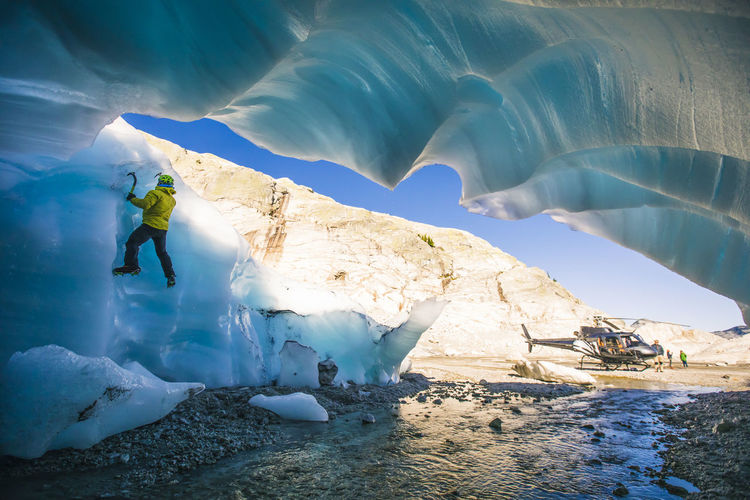 Man climbing on glacial ice next to helicopter during adventure tour.