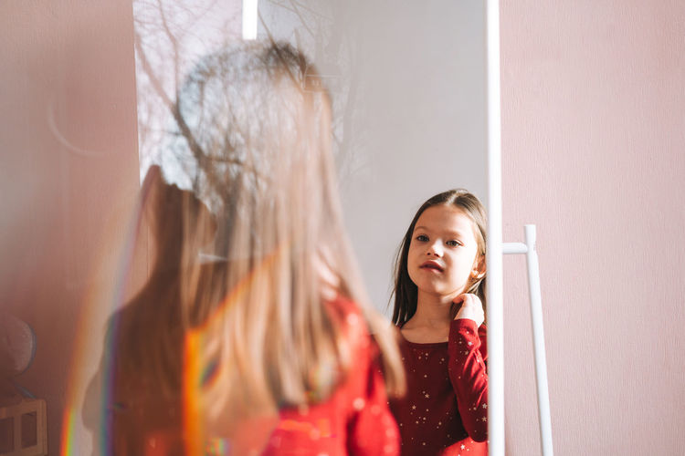 Cute tween girl with dark long hair in red dress looking in mirror in children room at cozy home