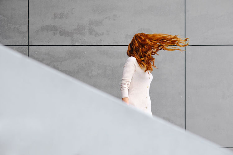 Side view of unrecognizable female with long red hair fluttering in wind standing against gray concrete wall on street