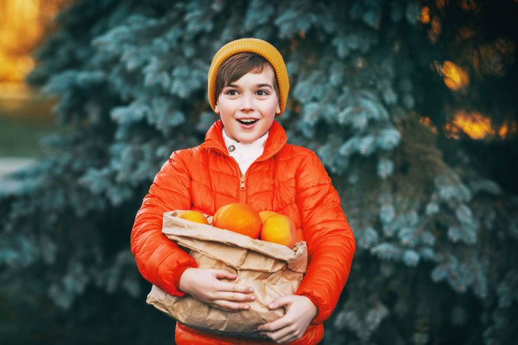 A boy in a bright orange jacket and a yellow hat holds a large package with oranges 