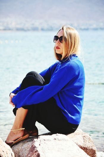 Woman wearing sunglasses sitting on rock by sea against sky
