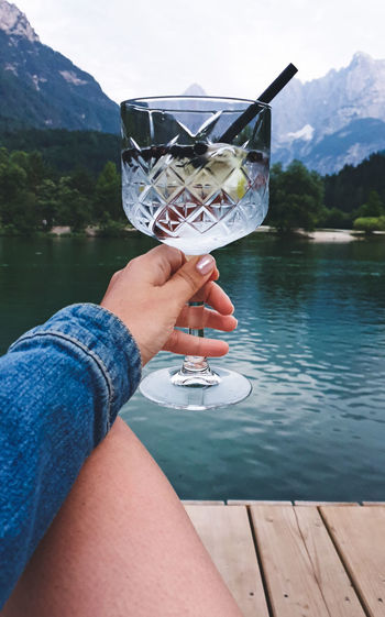 Personal perspective of woman holding glass with drink. lake, mountains in background.