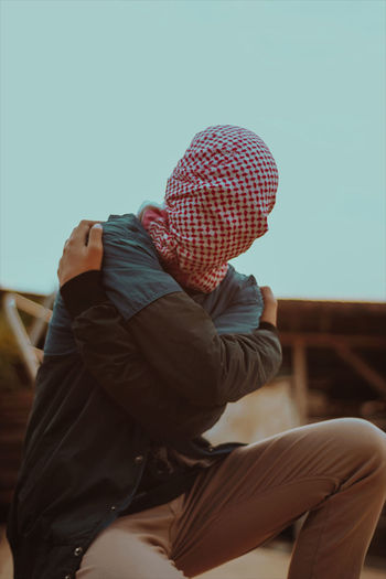 Man covering face with headscarf while crouching against clear sky