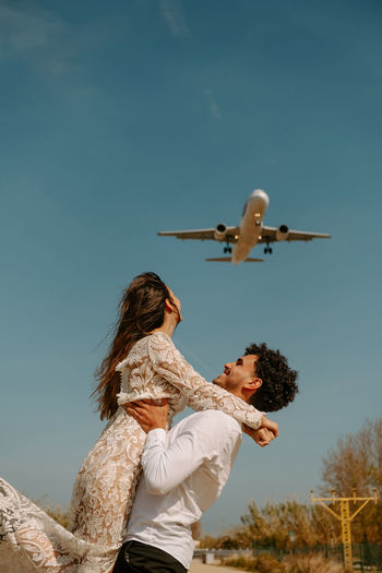 Side view of happy young man in stylish outfit carrying cheerful woman in white wedding dress while standing on country road against blue sky with flying airplane