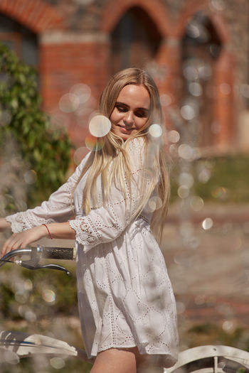 Pretty blond woman in white dress with bike near fountain in sunny light