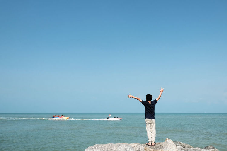 Rear view of boy standing on rock at beach against clear sky