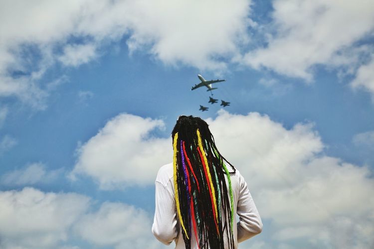 Rear view of man with colorful dreadlocks looking at airplanes flying against cloudy sky