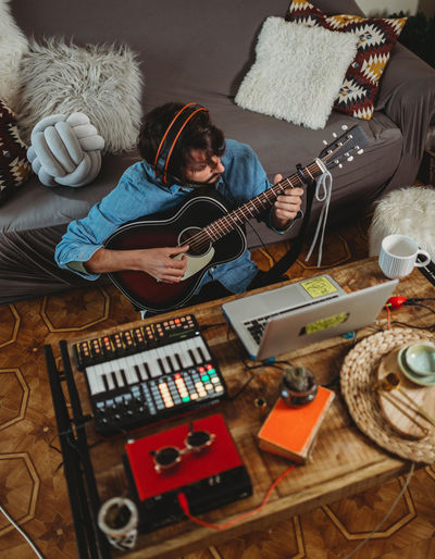 Young man in headphones playing on guitar near table with laptop and synthesizer at home looking at camera