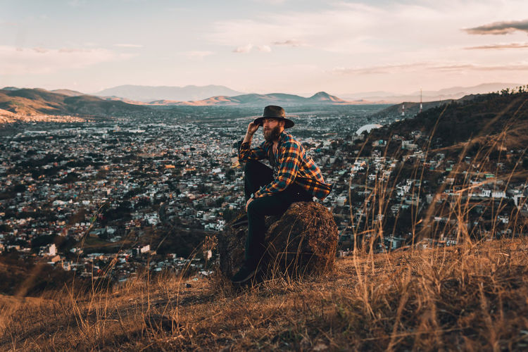 Male on rock posing in vintage clothes on golden hilltop overlooking a valley at sunset.
