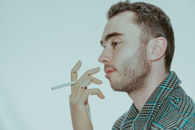 Side view of young man with robotic arm and cigarette looking away against white background