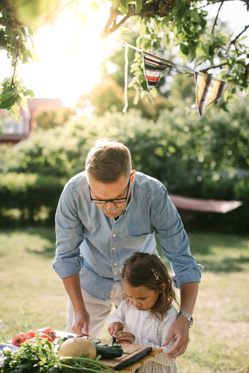 Grandfather assisting granddaughter in cutting vegetable at table in backyard