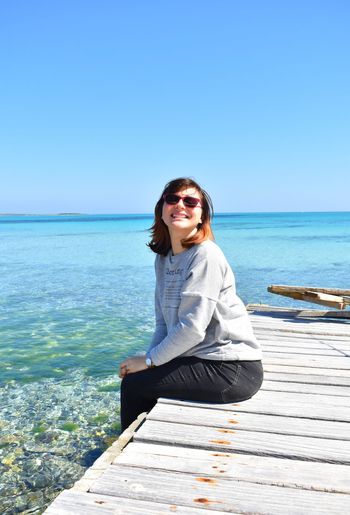 Young woman wearing sunglasses sitting on pier over sea against clear blue sky