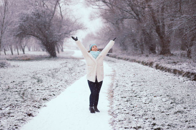 Happy young woman with arms raised standing at park during winter