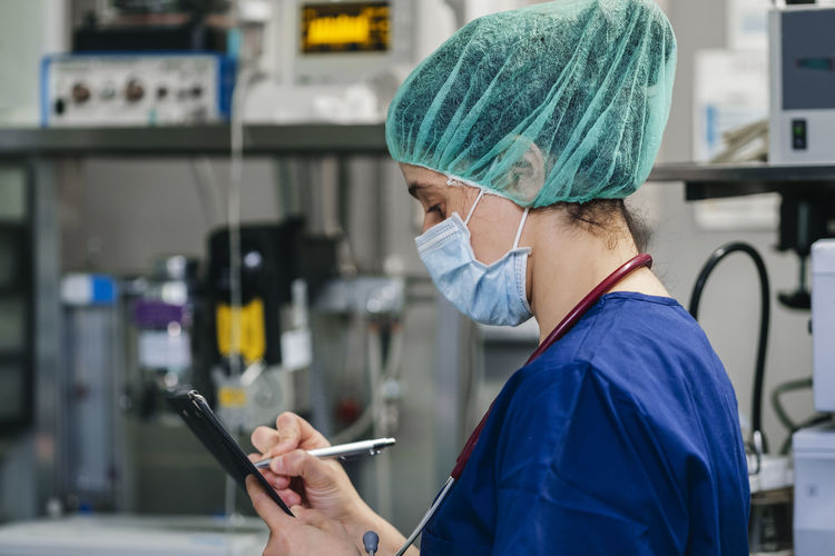 Side view of young female veterinary in medical mask and uniform standing in operating room and taking notes in clipboard with pen before surgery in clinic