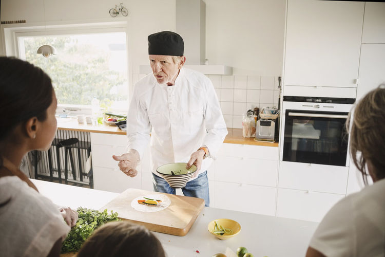 Senior man in chef's jacket talking to family while preparing food in kitchen