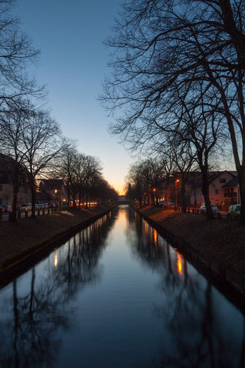 Canal amidst bare trees against sky at dusk