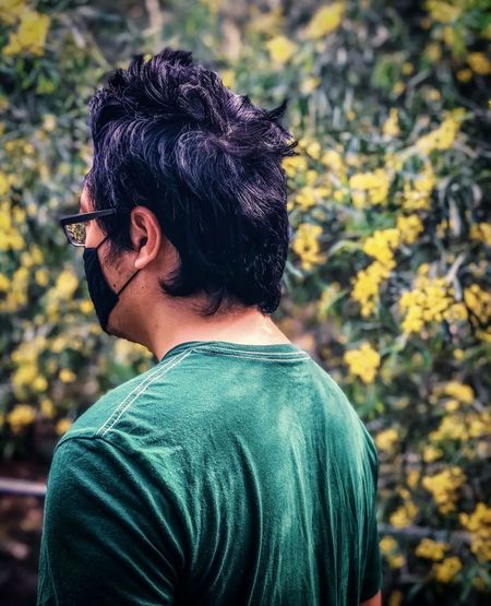 Rear view of young man wearing glasses and face mask against yellow flowering plants.