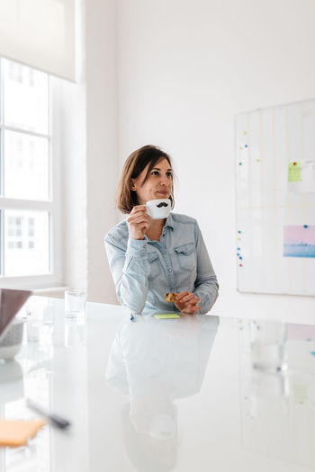 PORTRAIT OF WOMAN HOLDING SMART PHONE WHILE STANDING ON TABLE