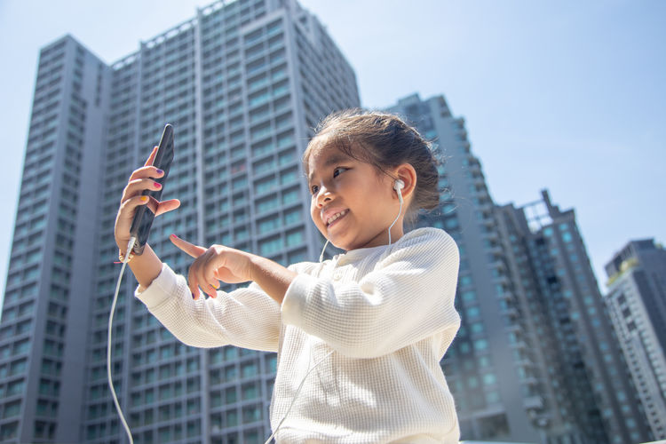 Low angle view of smiling girl using mobile phone while standing against buildings in city during sunny day