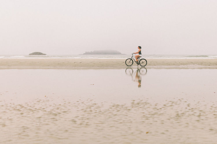 Side view of woman riding bicycle at beach against clear sky