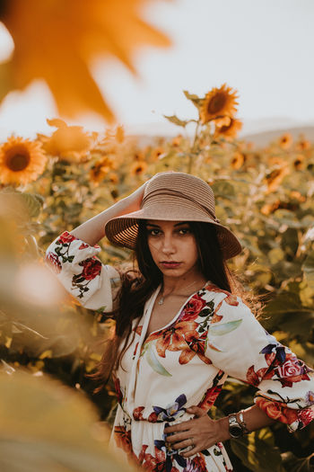 Portrait of young woman wearing hat standing against sunflowers