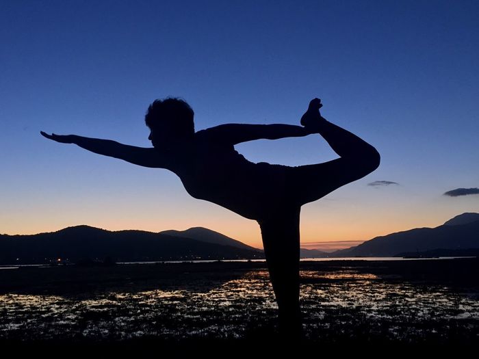 Silhouette man doing yoga at beach against clear sky during sunrise