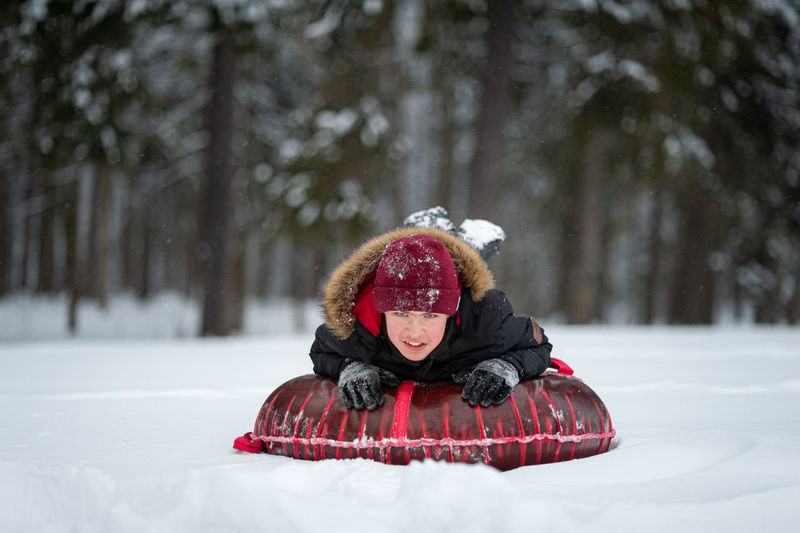 A smiling teenage boy in a black jacket with a fur and red hat lies on a tubing in a snow-covered