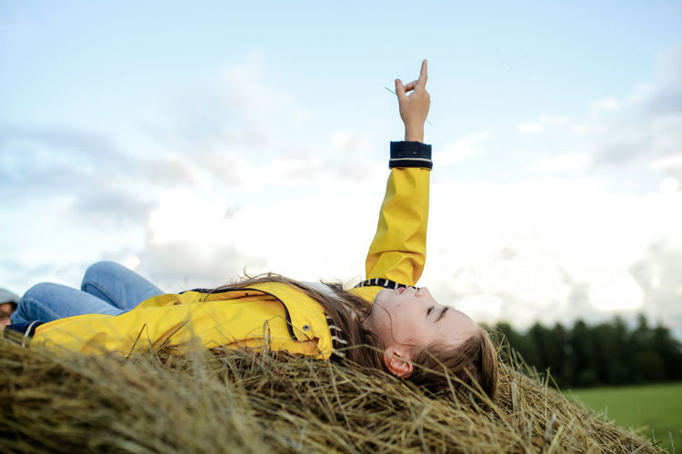 Girl pointing while lying on hay against sky