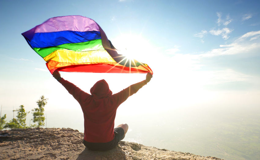 Rear view of man holding rainbow flag while sitting on mountain against sky