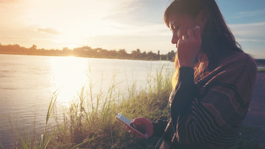 Young woman using phone by lake against sky during sunset