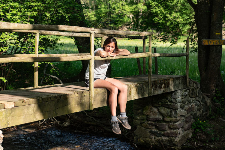 Calm female sitting on old bridge over river in woods and enjoying nature in summer